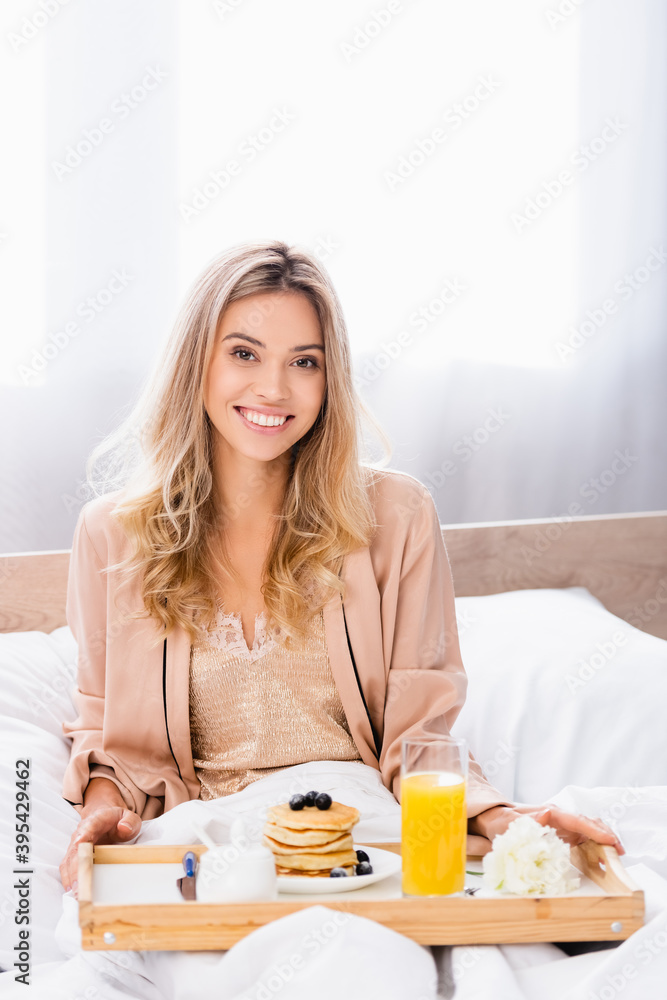  woman in pajamas holding tray with pancakes, orange juice and flower on bed