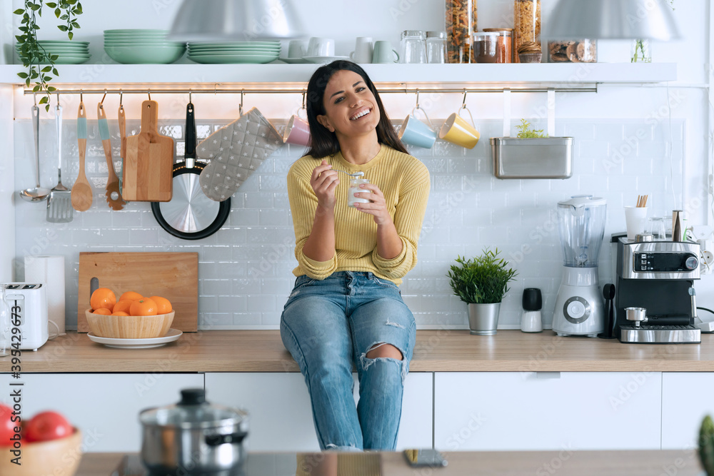 Pretty happy young woman eating yogurt while smiling thinking on her things sitting on the kitchen at home. Pretty happy young woman eating yogurt while smiling thinking on her things sitting on the kitchen at home.