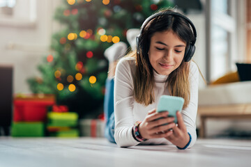 A young girl lying on the floor near the Christmas tree and using a mobile phone