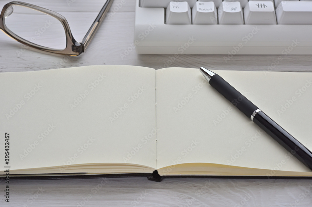 There's a keyboard, glasses, and an open white notebook on the desk. There's a keyboard, glasses, and an open white notebook on the desk.