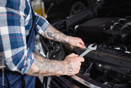 partial of tattooed technician holding wrench near car engine compartment on blurred foreground