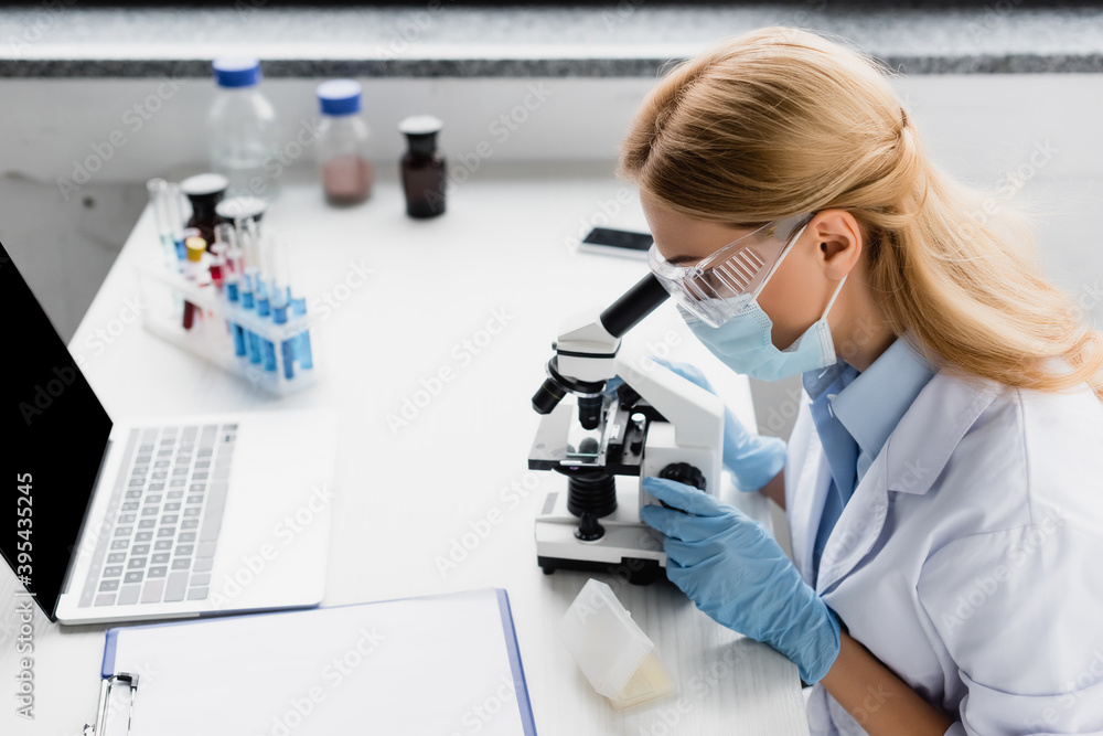 scientist in mask looking through microscope on desk scientist in mask looking through microscope on desk