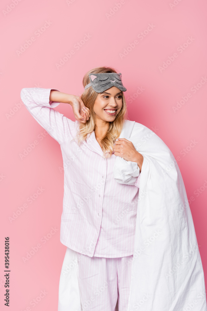 woman in pajamas and blindfold smiling while holding blanket on pink background woman in pajamas and blindfold smiling while holding blanket on pink background