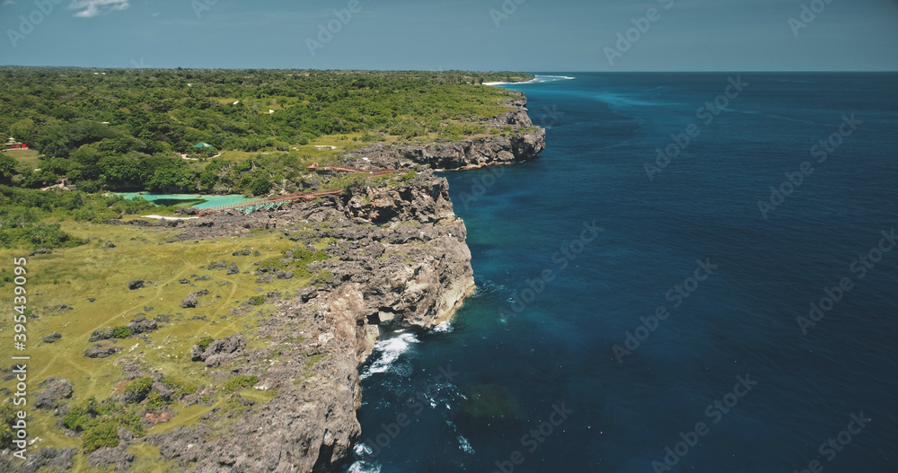 Cliff ocean coastline with green plants and trees at azure limpid lake. Indonesia attraction with building and bridge on rock sea shore of Sumba Island. Cinematic tropic landscape at drone shot