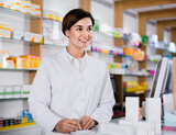 Young happy positive female pharmacist offering help in choosing at counter in pharmacy