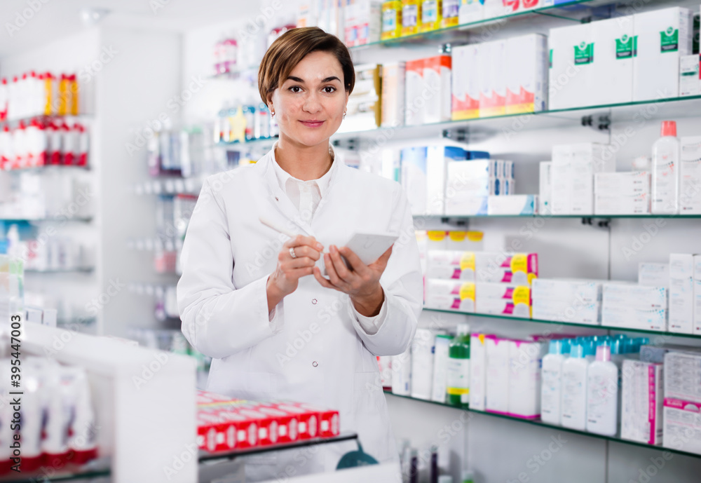 Diligent smiling female pharmacist noting assortment of drugs in pharmacy