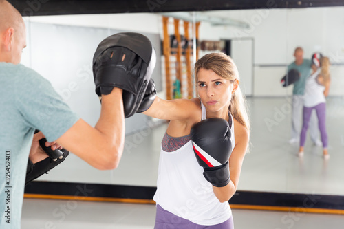 Portrait of young focused woman in boxing gloves launching blows on focus punch mitts during self defense workout in gym with trainer ..