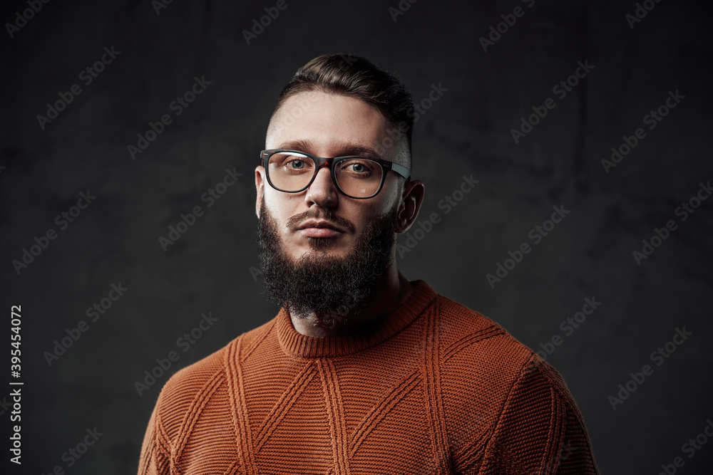 Portrait of a bearded and stylish guy with glasses he looks at camera with serious face and posing in dark background.