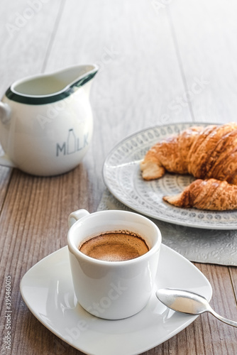vertical view of a white cup of espresso coffee with a small saucer with milk and a croissant on a dish on the background on the top of a wooden table