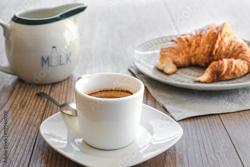 horizontal view of a white cup of espresso coffee with a small saucer with milk and a croissant on a dish on the background on the top of a wooden table