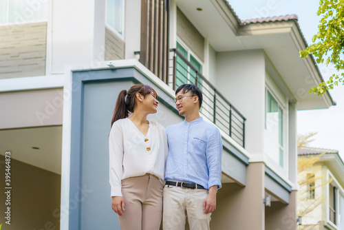 Portrait of Asian young couple standing and hugging together looking happy in front of their new house to start new life. Family, age, home, real estate and people concept.