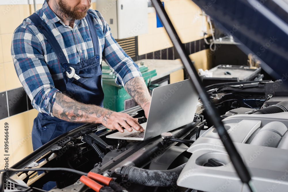 partial of tattooed repairman making car engine compartment diagnostic with laptop on blurred foreground partial of tattooed repairman making car engine compartment diagnostic with laptop on blurred foreground