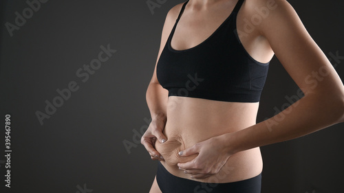 Woman in black underwear holding her fat belly, stomach with hands, diet, weight loss and liposuction concept. Isolated on grey background © Tatsiana