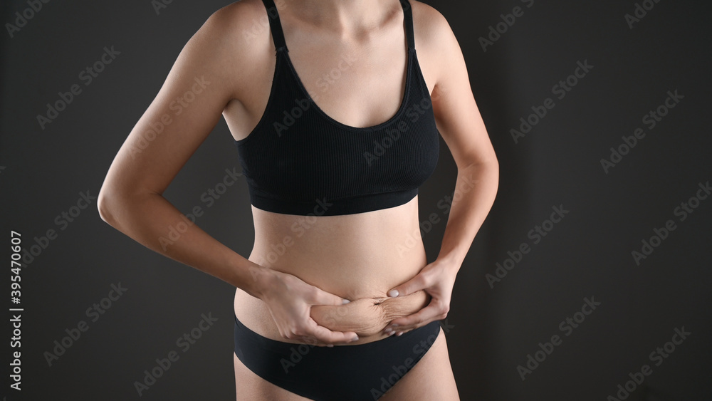 Fototapeta premium Woman in black underwear holding her fat belly, stomach with hands, diet, weight loss and liposuction concept. Isolated on grey background
