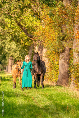 In the golden sun a portrait of a blond smiling girl with her horse and foal in the forest. Wearing a green dress. Selective focus © Dasya - Dasya