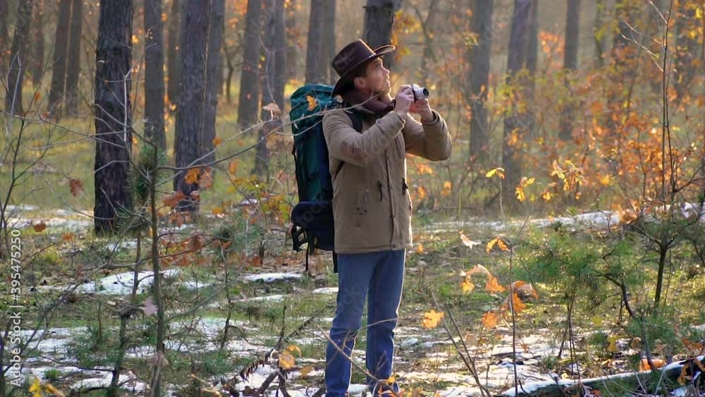 A man in a hat with a backpack takes pictures in the forest. Beautiful autumn forest and the first snow. Travel concept. Slow motion A man in a hat with a backpack takes pictures in the forest. Beautiful autumn forest and the first snow. Travel concept. Slow motion
