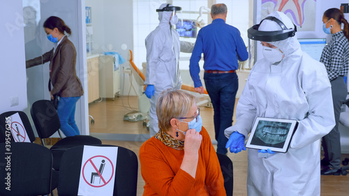 Dentist doctor with face shield pointing on tablet display explaining dental x-ray to senior patient during global pandemic. Nurse wearing protection suit  overall  mask and gloves  new normal