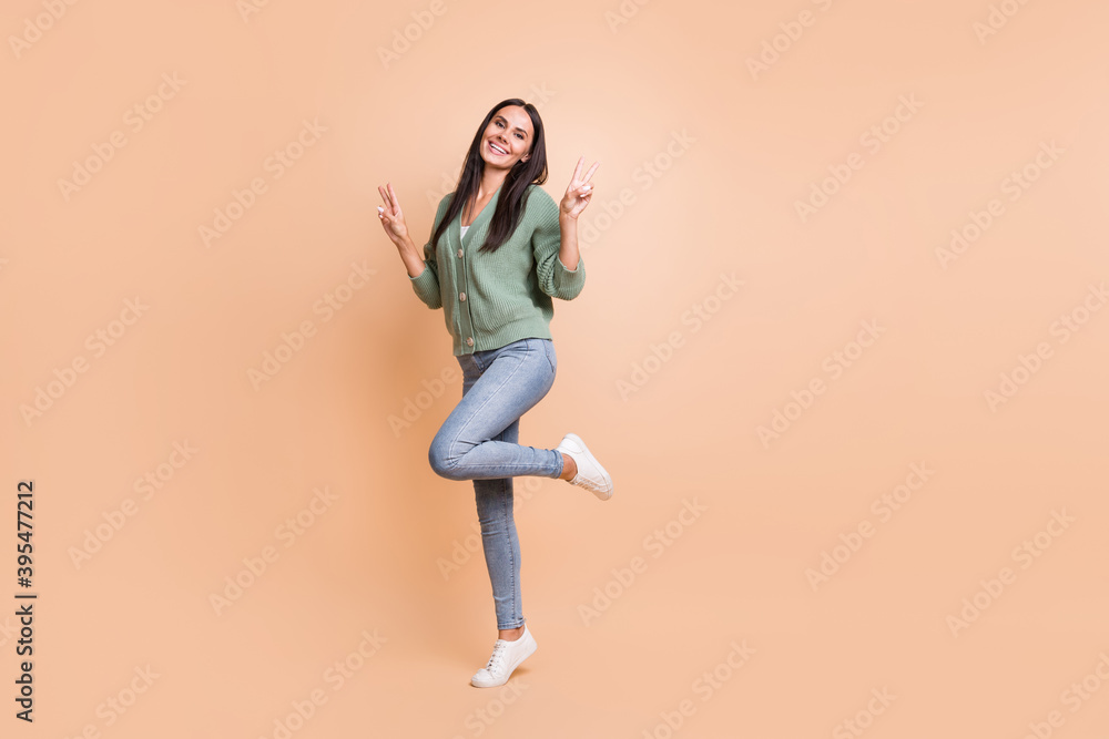 Full length photo of charming joyful woman show two v-signs hello stand one leg isolated on beige color background Full length photo of charming joyful woman show two v-signs hello stand one leg isolated on beige color background