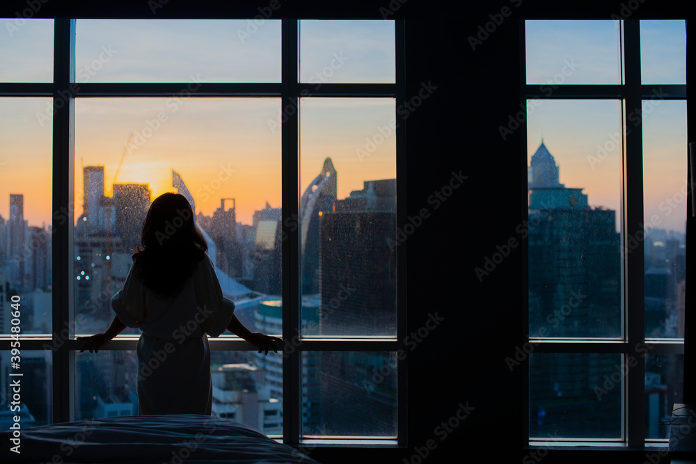 Fototapeta premium Young happy woman standing in the bedroom by the window with her back and beautiful sunrise or sunset and city scenery in the window. Copy space