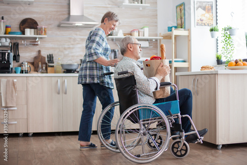 Cheerful wife helping disabled husband in the kitchen. Senior woman taking grocery paper bag from handicapped husband in wheelchair. Mature people with fresh vegetables from market. Living with