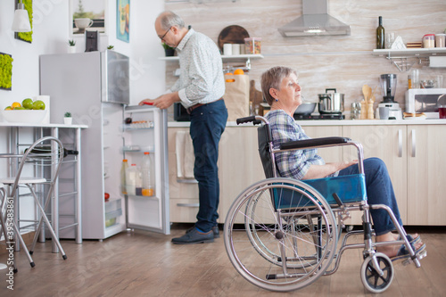 Senior man opening refrigerator while his disabled wife is sitting in wheelchair in kitchen looking through window. Living with handicapped person. Husband helping wife with disability. Elderly couple