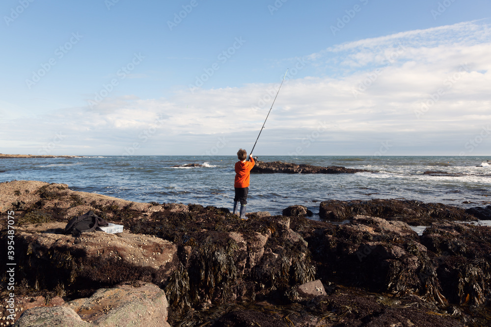 A 6yr old boy is fishing in the sea at Crail, Fife, Scotland. His tackle box and a bag are on the rocks behind him. He is using a weedless lure.