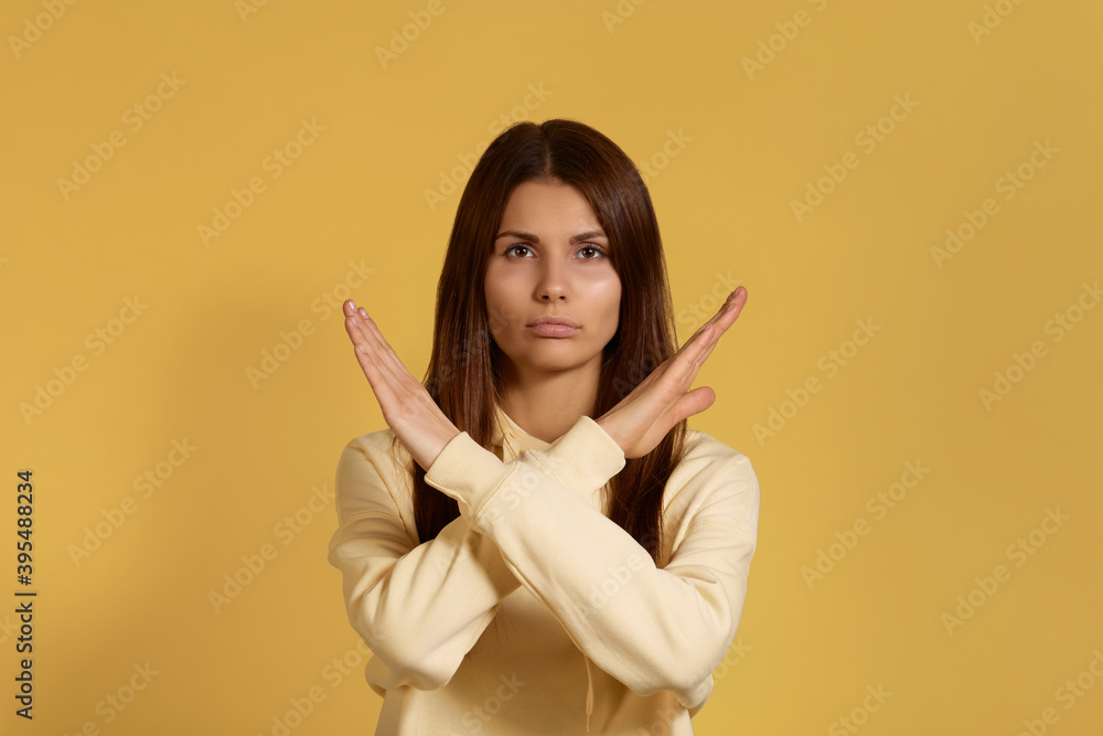 Cant go further. Serious caucasian woman in yellow hoodie keeps arms crossed, makes stop or ban gesture, indicates that the passage is closed. Isolated on a yellow background. Cant go further. Serious caucasian woman in yellow hoodie keeps arms crossed, makes stop or ban gesture, indicates that the passage is closed. Isolated on a yellow background.
