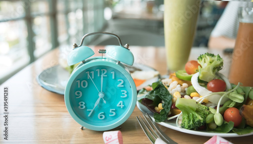Selective focus of Blue clock with woman eating salad bowl with fruit after intermittent fasting sitting on a table at home