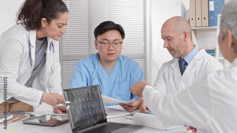 Over shoulder of unrecognizable grey-haired doctor looking at medical paper, passing in to young Asian male colleague across table. Multi Ethnic people talking, working in office Over shoulder of unrecognizable grey-haired doctor looking at medical paper, passing in to young Asian male colleague across table. Multi Ethnic people talking, working in office