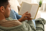 Man covered with warm green plaid reading book at home, closeup