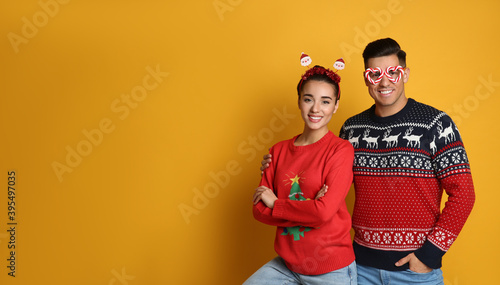 Couple in Christmas sweaters, Santa headband and party glasses on yellow background, space for text