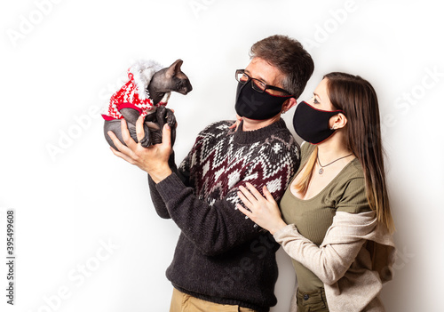 Couple in protective black face masks with a cat Peterbald in Christmas clothes during covd quarantine. Coronavirus new year concept on white background. New normal. Quarantine celebration photo