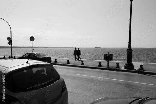two older men go for a stroll down the pathway walkway by the water sea in black and white one is wearing a hat by the road with parked cars