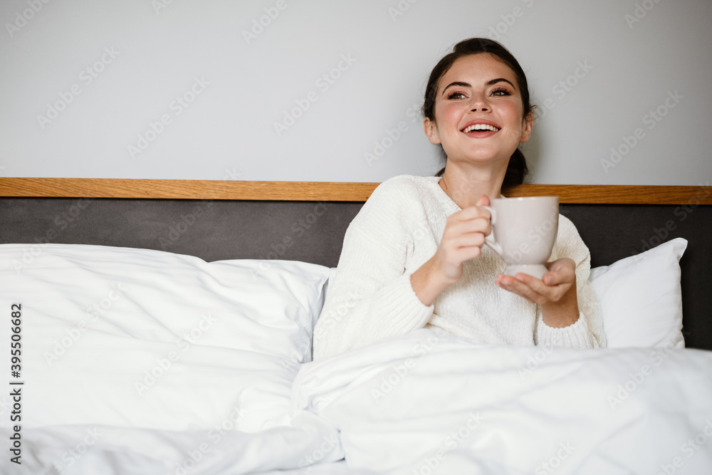 Charming cheerful girl smiling and drinking coffee while sitting in bad Charming cheerful girl smiling and drinking coffee while sitting in bad