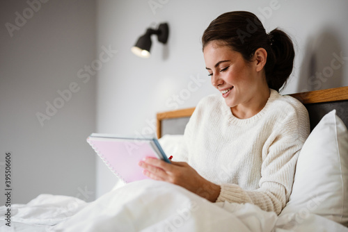 Charming cheerful girl writing down notes while sitting in bad