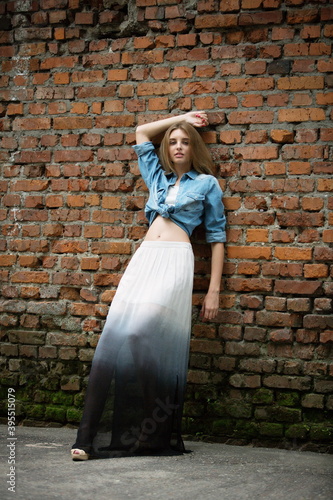 beautiful slender blonde girl in a long skirt and short denim jacket near an old brick wall