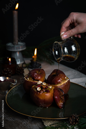 Pouring syrup on one of three baked apples stuffed with nuts, raisins and lingonberries on big green plate  on table decorated with candles, cones, fir branches on dark background .