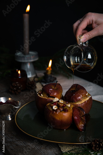 Pouring syrup on one of three baked apples stuffed with nuts, raisins and lingonberries on big green plate on table decorated with candles, cones, fir branches on dark background .