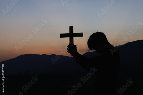 Silhouette of human kneeling down praying and holding christian cross for worshipping God at sunset background. Christian, Christianity, Religion copy space background.