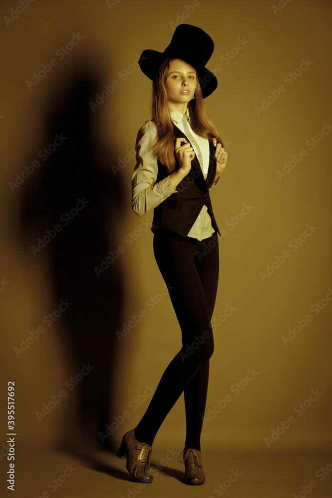 beautiful slender long-legged blonde model in leggings, white shirt, vest and a large vintage hat posing in the studio. the image is toned in olive color.