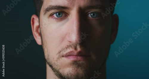 Cinematic close up of serious self confident young man with blue eyes is looking and blinking in camera isolated on dark background.