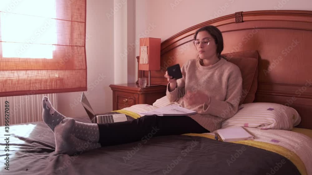 Young female student using her phone lying on bed, studying at home. Stay at home concept. Young female student using her phone lying on bed, studying at home. Stay at home concept.