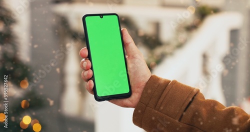 Fotografie Close up shot of black smartphone with green screen in Caucasian male hand while standing outdoors on Christmas Eve