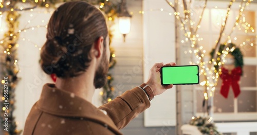 Obraz na plátně Close up shot of black smartphone with green screen in Caucasian male hand while standing outdoors on Christmas Eve