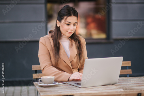Young business woman sitting on terrace by cafe with laptop. Woman dring coffee and working outdoors. Female use phone and notebook © Aleksandr