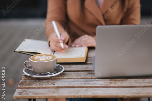 Close up of working proces in cafe outdoors. Woman writing down something in notebook. Cappuccino laptop and calendar on table