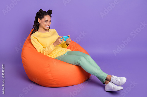 Full size photo of optimistic girl lying on pouf play telephone wear yellow shirt trousers sneakers isolated on lilac color background