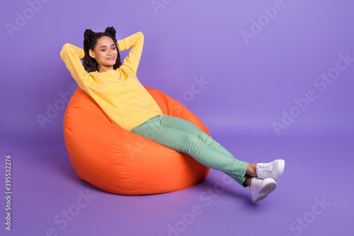 Full size photo of optimistic girl lying on pouf wear yellow shirt trousers sneakers isolated on lilac color background