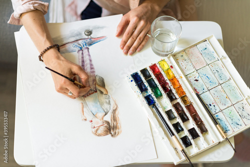 Close-up, hands of a woman artist, view from the top, a girl on a skateboard draws a picture, a color palette on the table, a glass cup with water background.