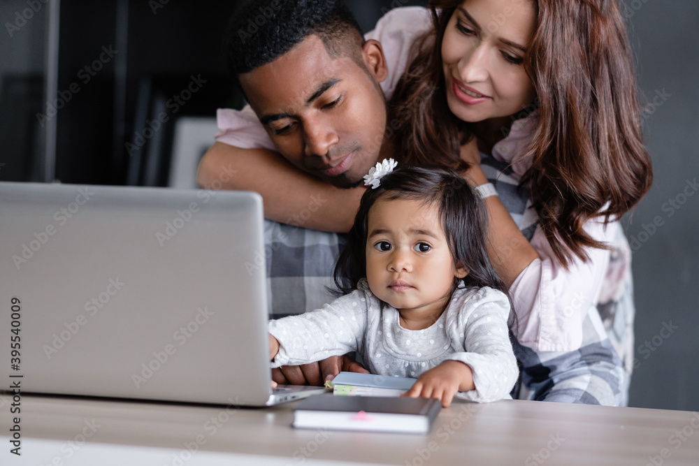 Happy friendly mixed race a family african-american father asian mother and charming daughter are sitting at a table with a laptop and a notebook. Freelance work and happy family concept